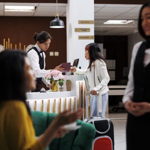 Male front desk receptionist at luxury resort checking visitors passports before registration. Asian visitor sipping coffee in modern waiting lounge ready to make reservation at modern hotel